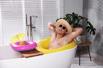 Photo of Happy man in sunglasses and straw hat with inflatable ring chilling in bathtub at home. Dreaming about vacation at sea