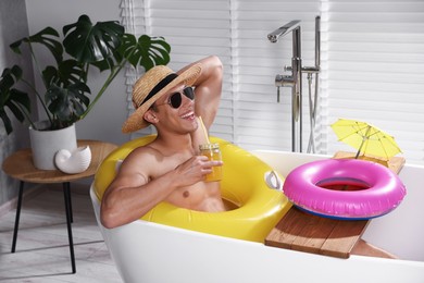 Photo of Happy man in sunglasses and straw hat drinking juice from mason jar with inflatable ring in bathtub at home. Dreaming about vacation at sea