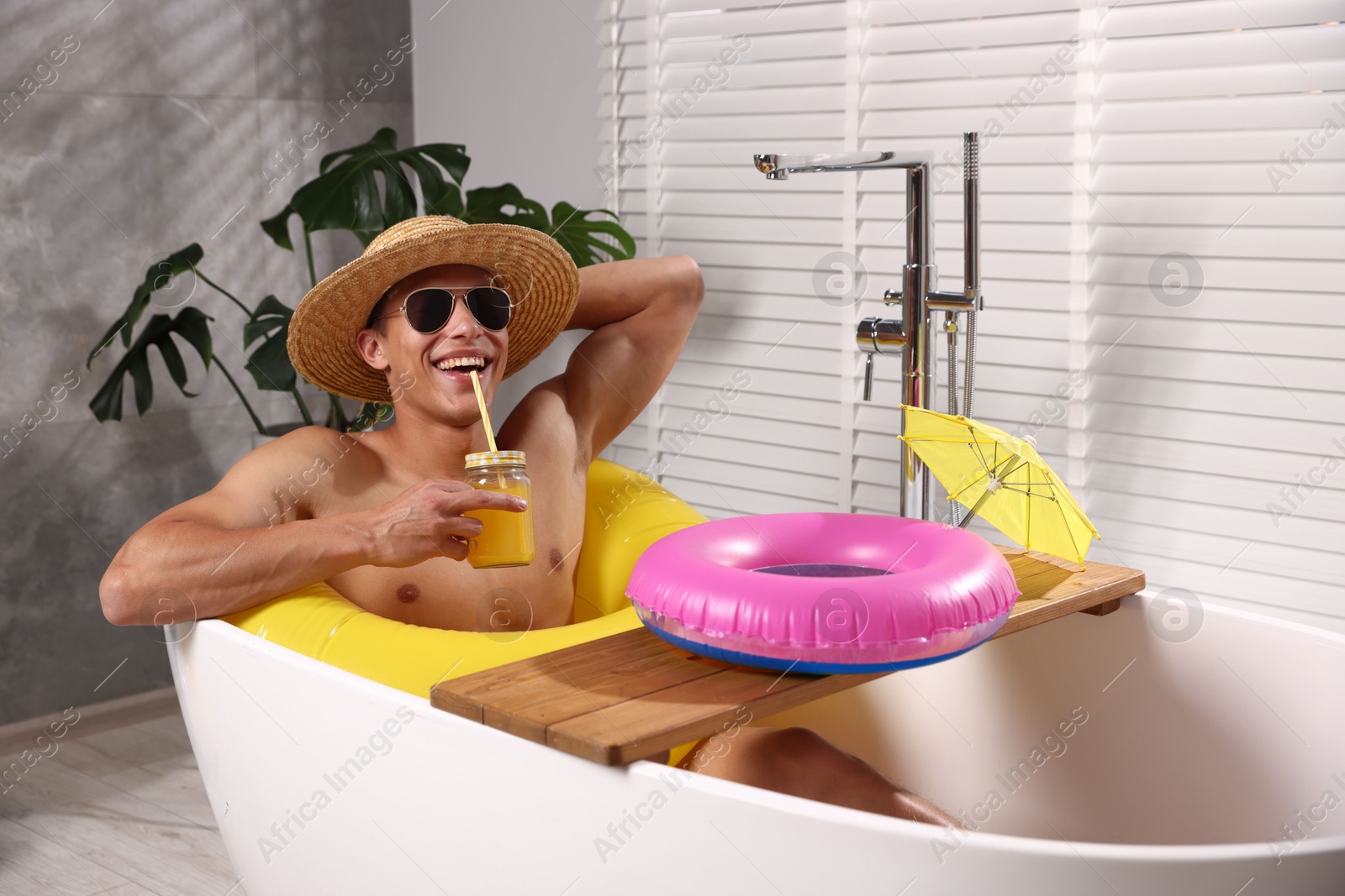 Photo of Happy man in sunglasses and straw hat drinking juice from mason jar with inflatable ring in bathtub at home. Dreaming about vacation at sea