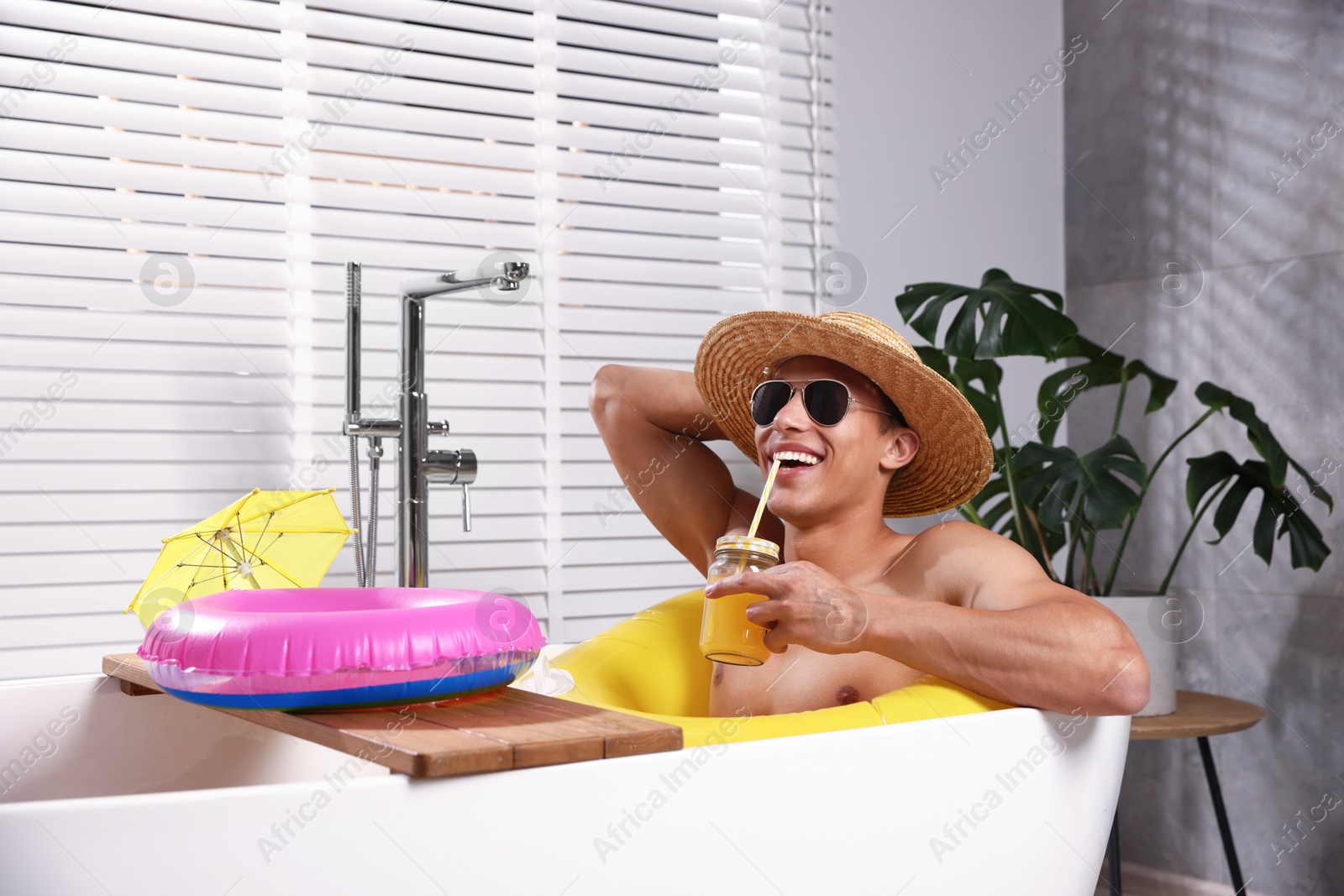 Happy man in sunglasses and straw hat drinking juice from mason jar with inflatable ring in bathtub at home. Dreaming about vacation at sea Photo of Happy man in sunglasses and straw hat drinking juice from mason jar with inflatable ring in bathtub at home. Dreaming about vacation at sea