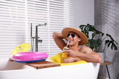 Photo of Happy man in sunglasses and straw hat drinking juice from mason jar with inflatable ring in bathtub at home. Dreaming about vacation at sea