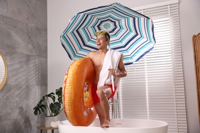 Photo of Happy man in with inflatable ring, diving mask, towel and beach umbrella in bathtub at home. Dreaming about vacation at sea