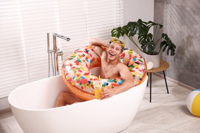 Photo of Happy man with diving mask, mason jar of juice and inflatable ring chilling in bathtub at home. Dreaming about vacation at sea