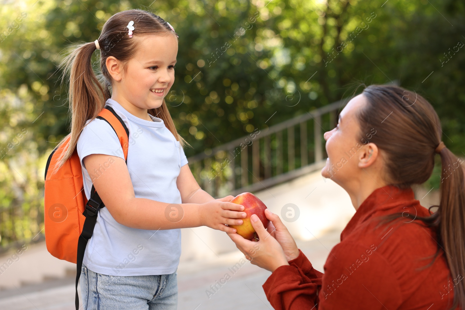 Mother giving apple to her daughter while taking to school outdoors Photo of Mother giving apple to her daughter while taking to school outdoors