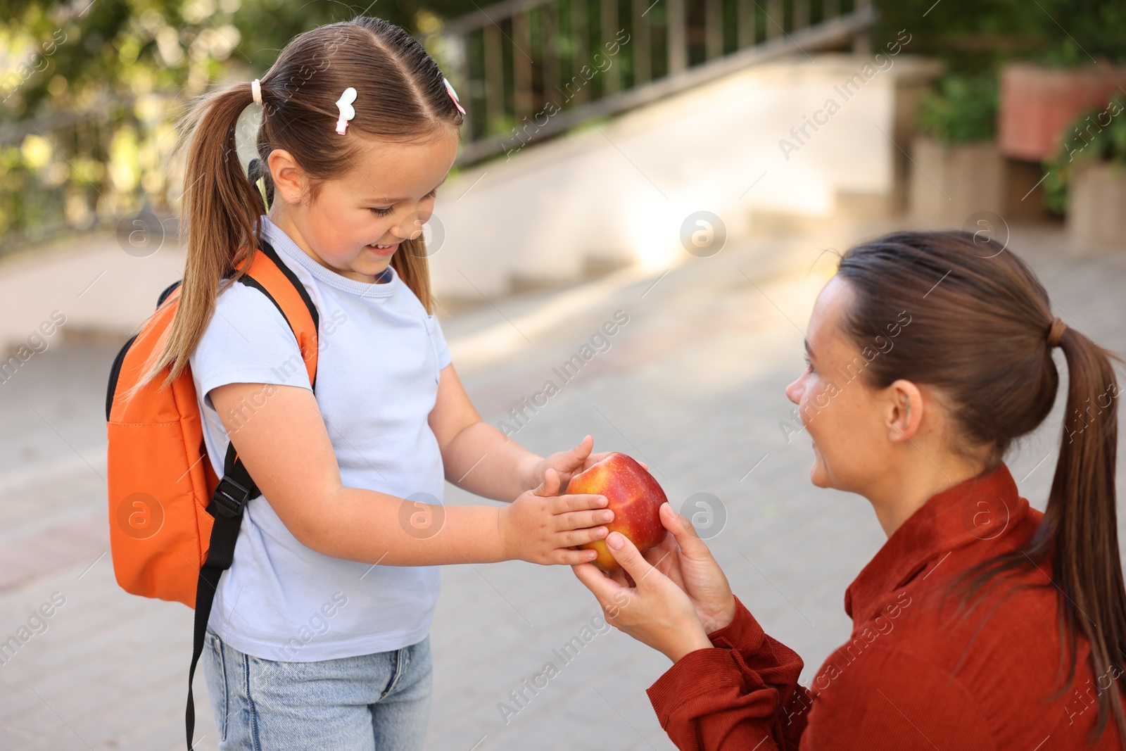 Mother giving apple to her daughter while taking to school outdoors Photo of Mother giving apple to her daughter while taking to school outdoors