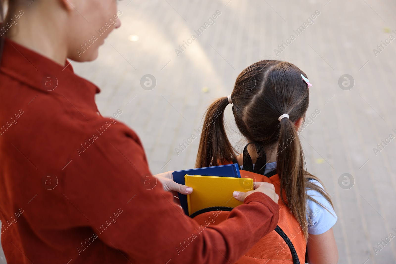 Photo of Mother putting books into her daughter's backpack while taking her to school outdoors, closeup