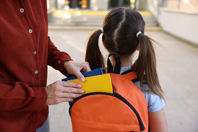 Photo of Mother putting books into her daughter's backpack while taking her to school outdoors, closeup