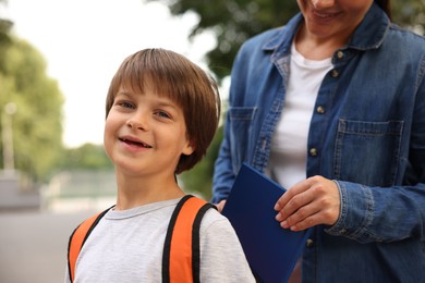 Photo of Mother putting book into son's backpack while taking him to school outdoors, closeup