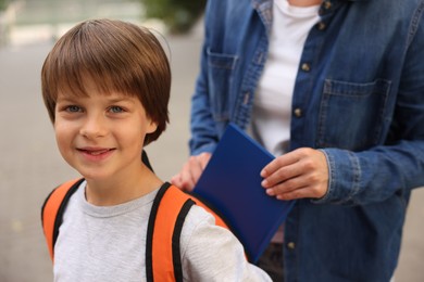 Photo of Mother putting book into son's backpack while taking him to school outdoors, closeup