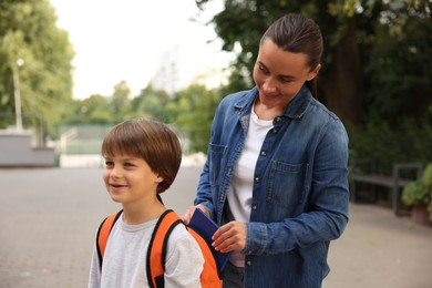Photo of Mother putting book into son's backpack while taking him to school outdoors