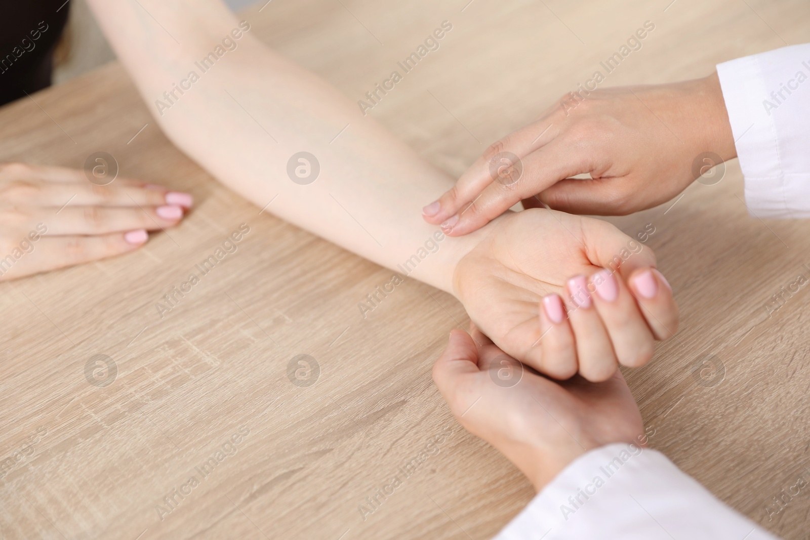 Photo of Doctor checking woman's pulse with fingers at wooden table indoors, closeup