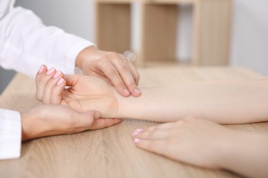Photo of Doctor checking woman's pulse with fingers at wooden table indoors, closeup