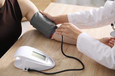 Photo of Doctor checking woman's heart rate and blood pressure via sphygmomanometer at wooden table indoors, closeup