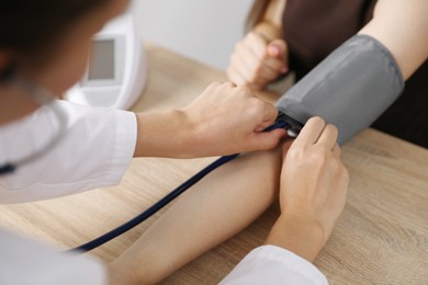 Photo of Doctor checking woman's heart rate and blood pressure via sphygmomanometer at wooden table indoors, closeup