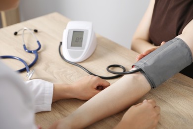 Photo of Doctor checking woman's heart rate and blood pressure via sphygmomanometer at wooden table indoors, closeup