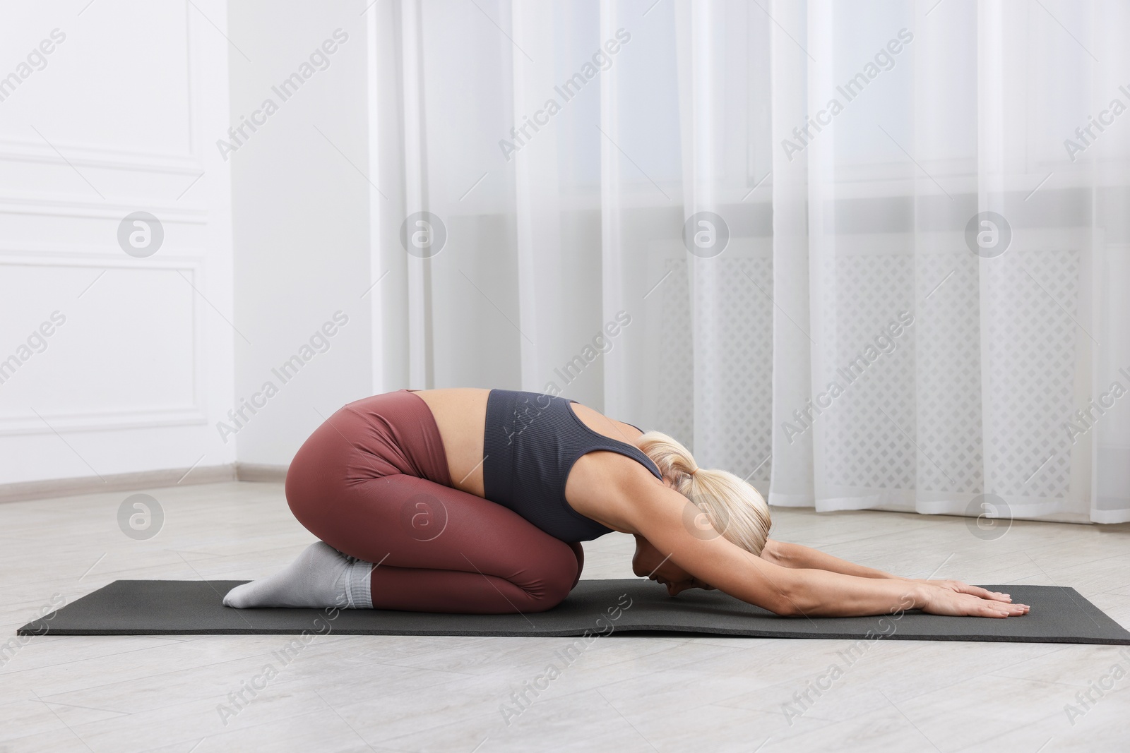 Photo of Mature woman in sportswear stretching on mat near window at home