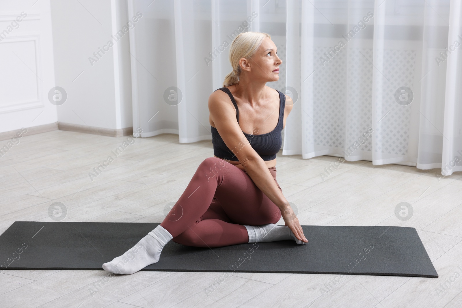 Photo of Mature woman in sportswear stretching on mat near window at home