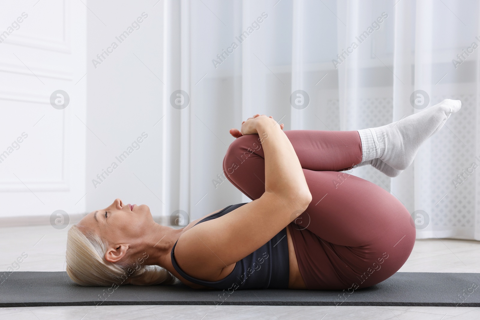 Photo of Mature woman in sportswear stretching on mat near window at home