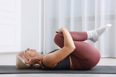 Photo of Mature woman in sportswear stretching on mat near window at home