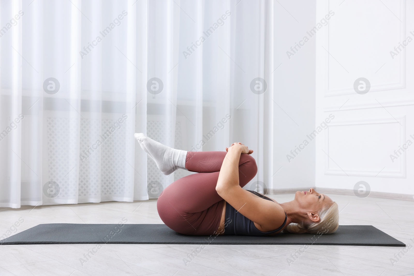 Photo of Mature woman in sportswear stretching on mat near window at home