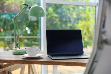 Photo of Home workspace. Laptop, lamp and cup of drink on wooden desk near window indoors