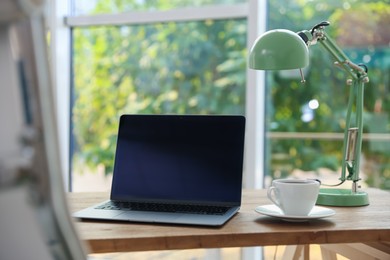 Photo of Home workspace. Laptop, lamp and cup of drink on wooden desk near window indoors