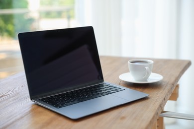 Photo of Home workspace. Laptop and cup of drink on wooden desk near window indoors, closeup