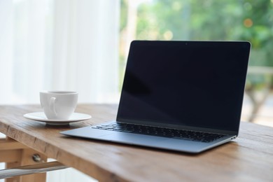 Photo of Home workspace. Laptop and cup of drink on wooden desk near window indoors, closeup