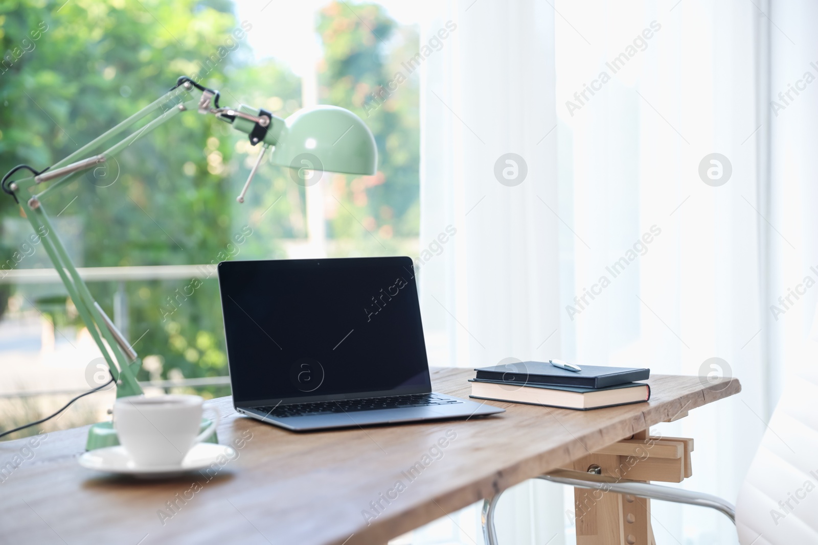 Photo of Home workspace. Laptop, cup of drink, lamp and stationery on wooden desk near window indoors