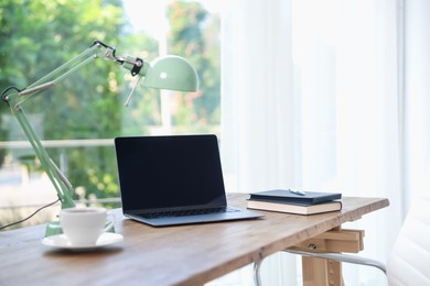 Photo of Home workspace. Laptop, cup of drink, lamp and stationery on wooden desk near window indoors