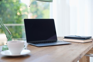 Photo of Home workspace. Laptop, cup of drink, lamp and stationery on wooden desk near window indoors