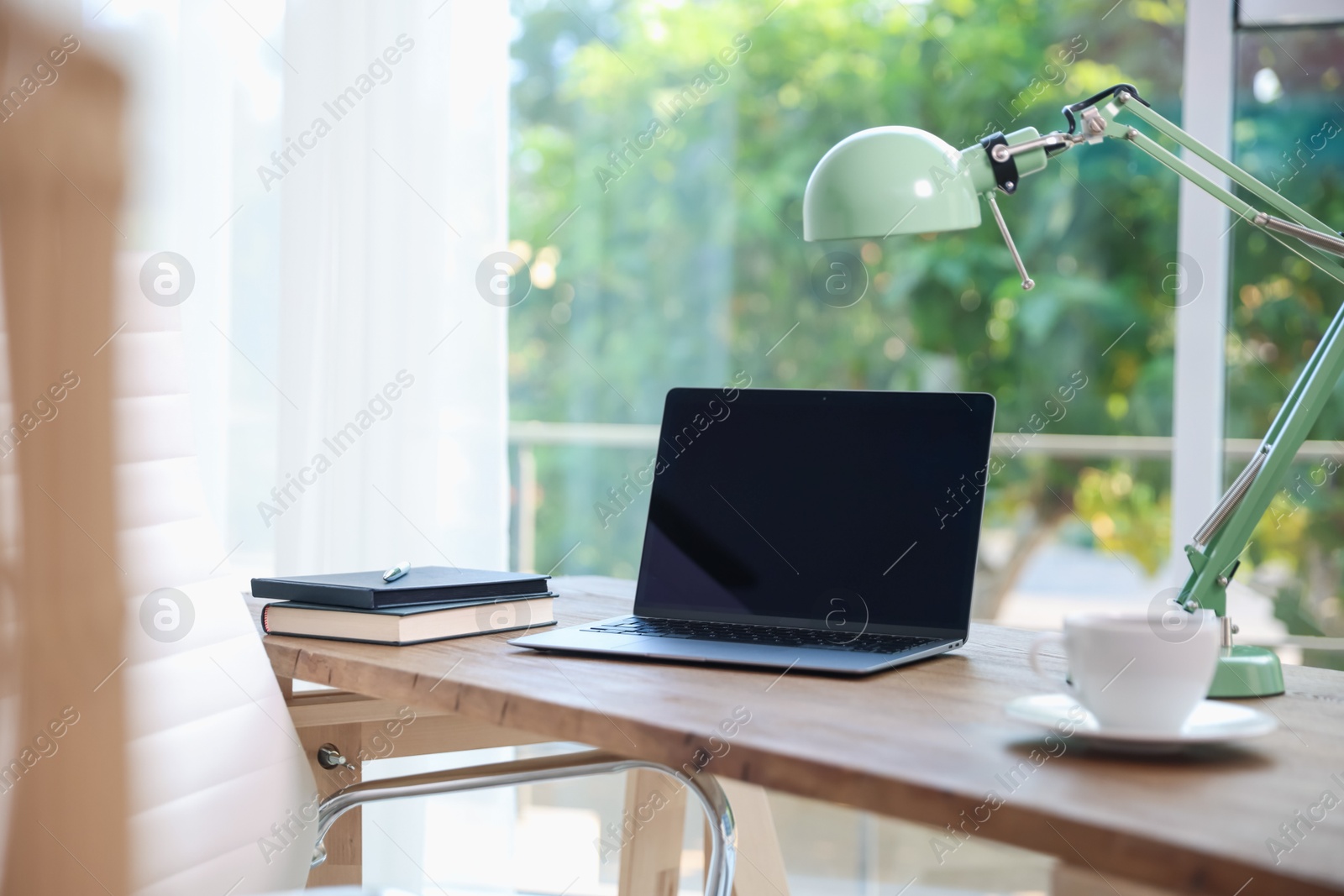 Photo of Home workspace. Laptop, cup of drink, lamp and stationery on wooden desk near window indoors