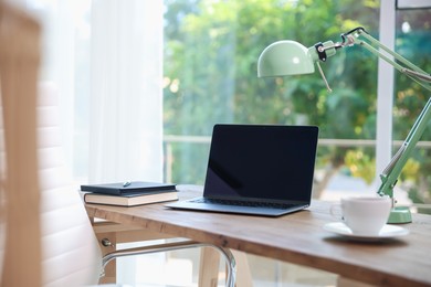 Photo of Home workspace. Laptop, cup of drink, lamp and stationery on wooden desk near window indoors