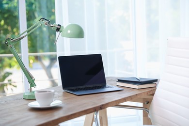 Photo of Home workspace. Laptop, cup of drink, lamp and stationery on wooden desk near window indoors
