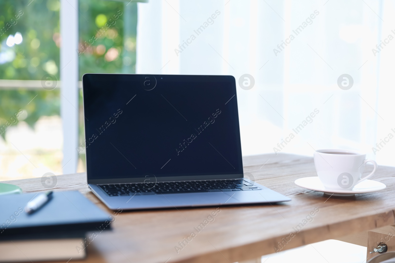 Photo of Home workspace. Laptop, cup of drink and stationery on wooden desk near window indoors