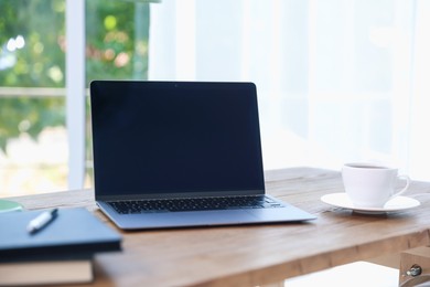 Photo of Home workspace. Laptop, cup of drink and stationery on wooden desk near window indoors