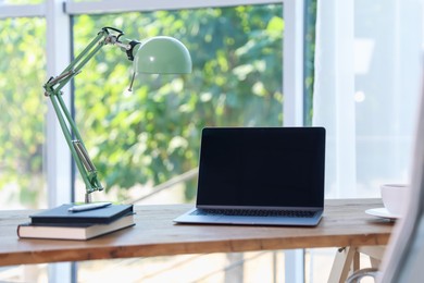 Photo of Home workspace. Laptop, cup of drink, lamp and stationery on wooden desk near window indoors