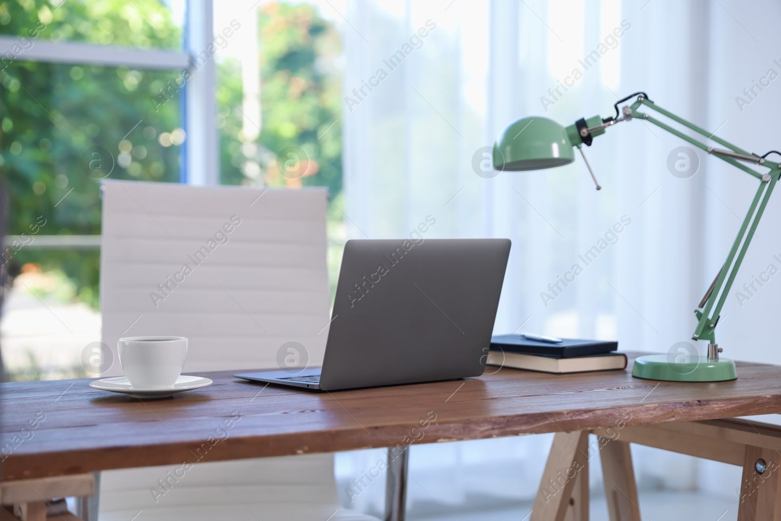 Photo of Home workspace. Laptop, cup of drink, lamp and stationery on wooden desk near window indoors