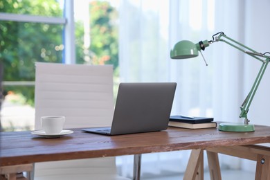 Photo of Home workspace. Laptop, cup of drink, lamp and stationery on wooden desk near window indoors