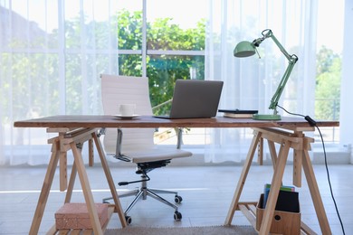 Photo of Home workspace. Laptop, cup of drink, lamp and stationery on wooden desk near window indoors