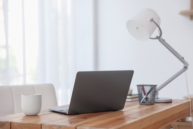 Photo of Home workspace. Laptop, cup of drink, lamp and stationery on wooden desk near window indoors