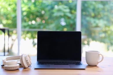 Photo of Home workspace. Laptop, cup of drink and headphones on wooden desk near window indoors