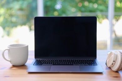 Photo of Home workspace. Laptop, cup of drink and headphones on wooden desk near window indoors