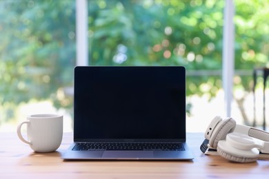 Photo of Home workspace. Laptop, cup of drink and headphones on wooden desk near window indoors