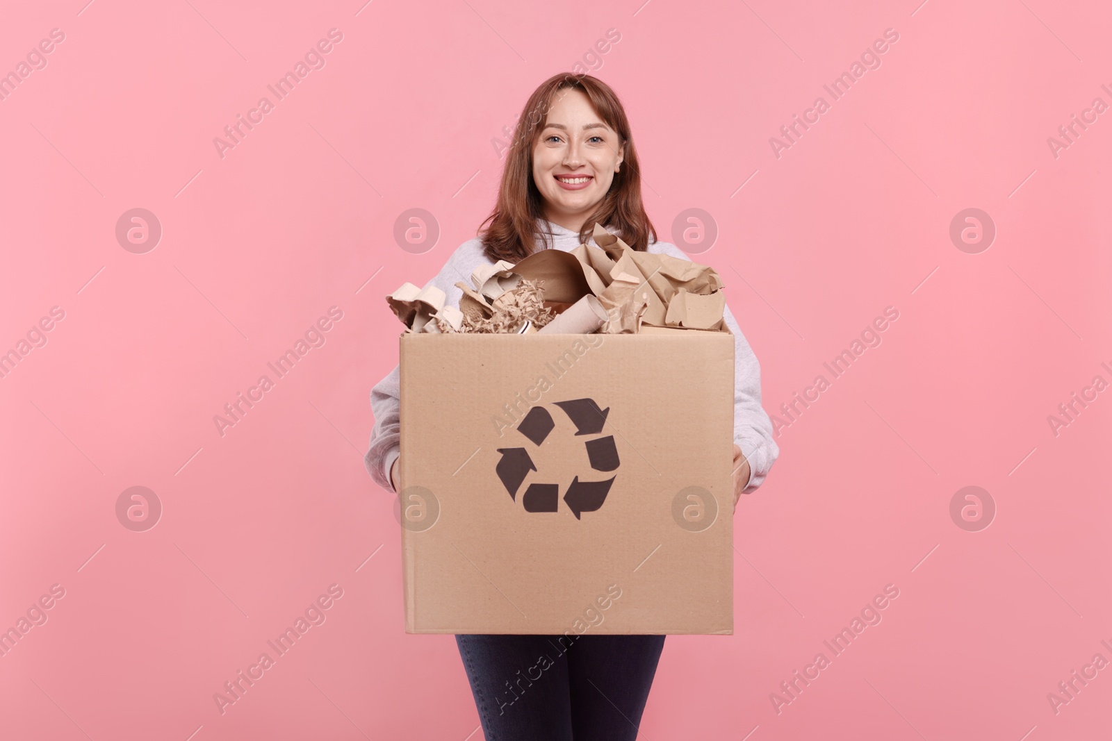 Recycling. Smiling woman holding cardboard box with different waste paper on pink background Photo of Recycling. Smiling woman holding cardboard box with different waste paper on pink background