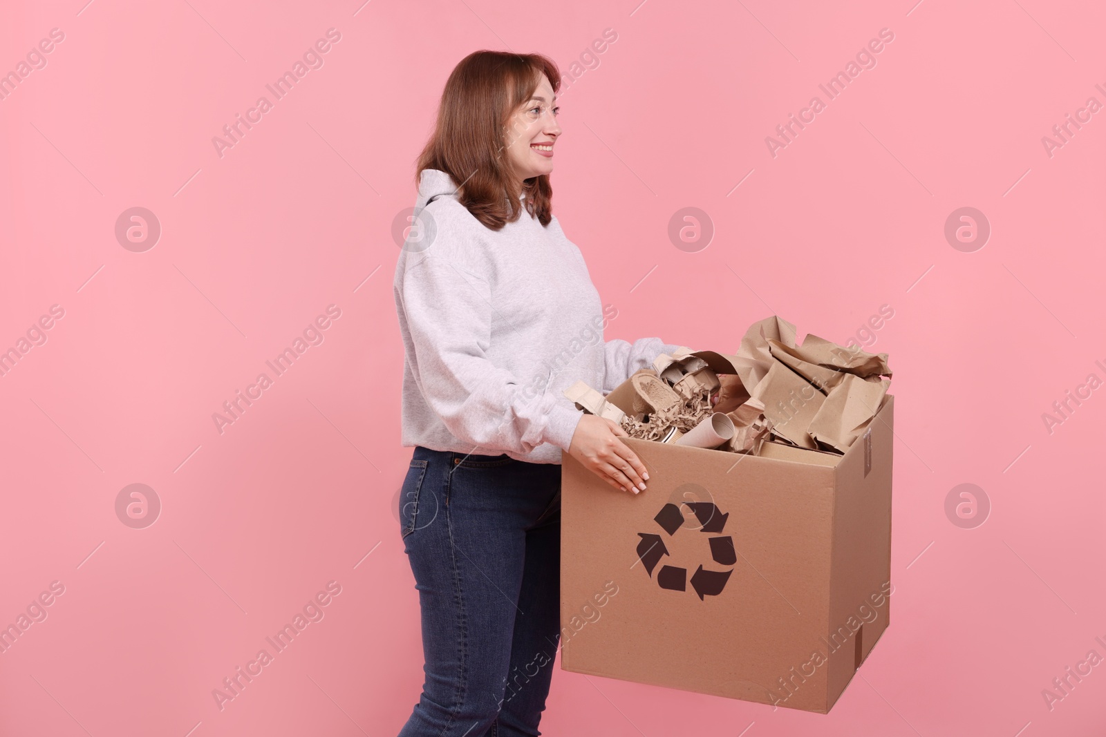 Recycling. Smiling woman holding cardboard box with different waste paper on pink background Photo of Recycling. Smiling woman holding cardboard box with different waste paper on pink background