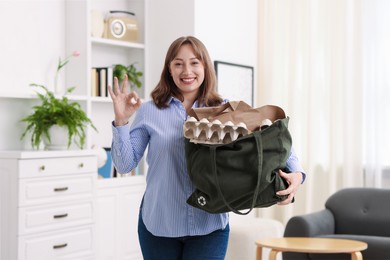 Photo of Recycling. Smiling woman with bag full of different waste paper showing OK gesture indoors