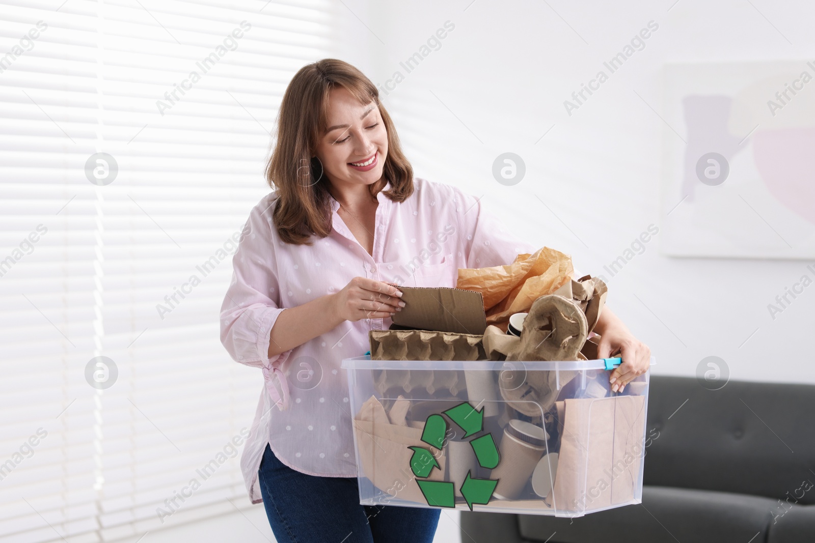 Photo of Recycling. Smiling woman holding plastic box with different waste paper indoors