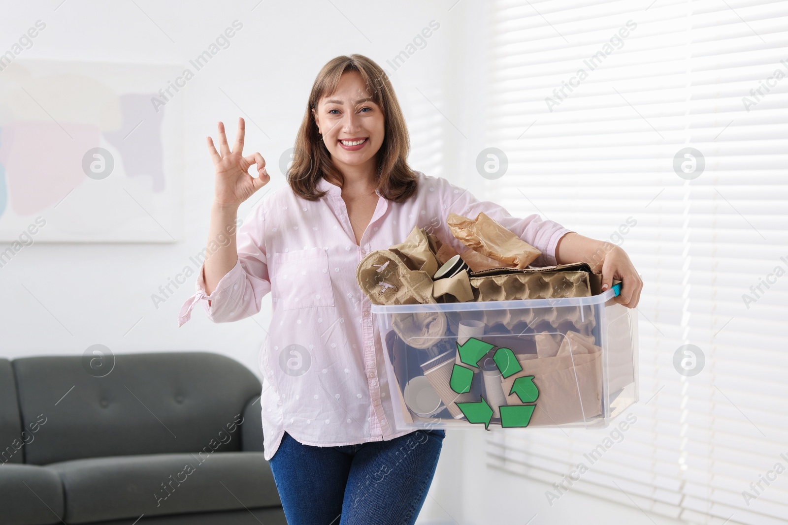 Photo of Recycling. Smiling woman holding plastic box with different waste paper and showing OK gesture indoors