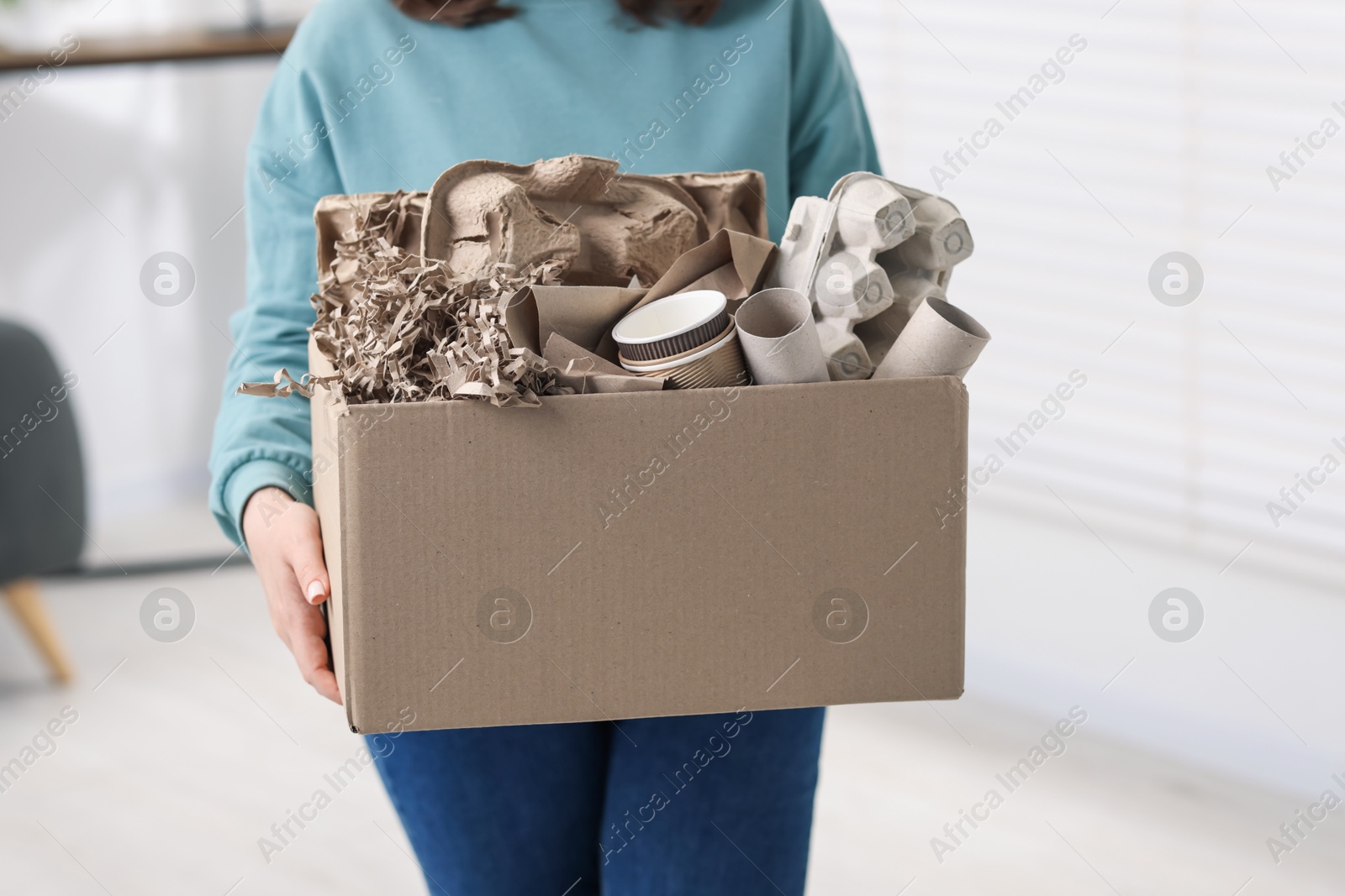 Photo of Recycling. Woman holding cardboard box with different waste paper indoors, closeup